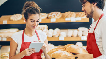Two bakers standing in front of shelves of bread while using a tablet to manage their business' online banking.