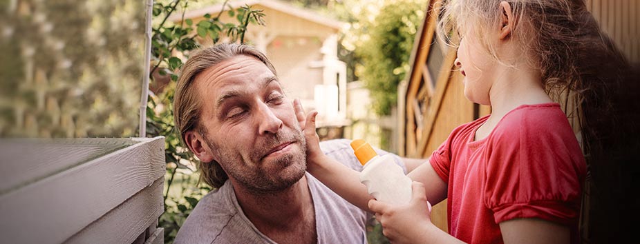 Image of man closing his eyes and smiling as his young daughter applies sunscreen to his face