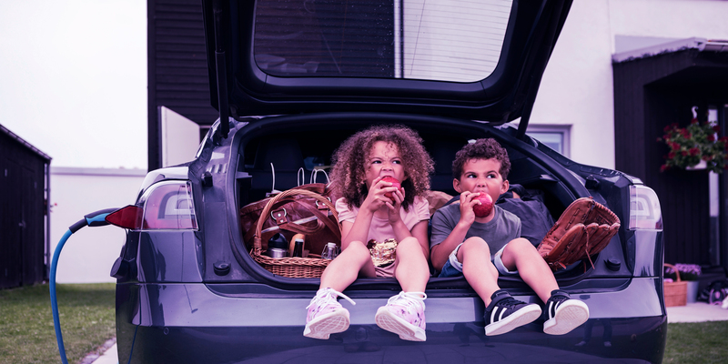 Children enjoying an apple in the boot of a parked electric car with picnic items, charging in a suburban neighborhood.